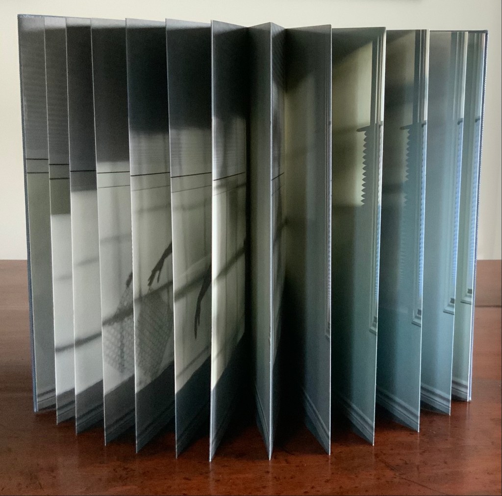 A side view of an artist's book with multiple accordion-fold pages, displaying layered photographs of shadows and light cast by windows and objects inside a room.