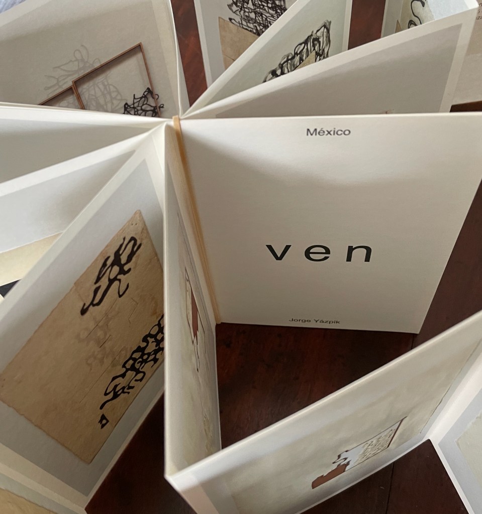 An open art book titled 'ven' by Jorge Yazpik featuring abstract designs and illustrations, displayed in a fan-like arrangement on a wooden surface.