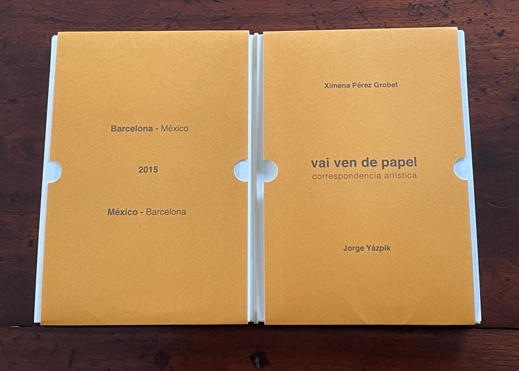 A photo of a closed book with an orange cover featuring the titles 'Barcelona - México' and 'vai ven de papel' along with the names Ximena Pérez Grobet and Jorge Yázpik. The book is displayed on a wooden surface.