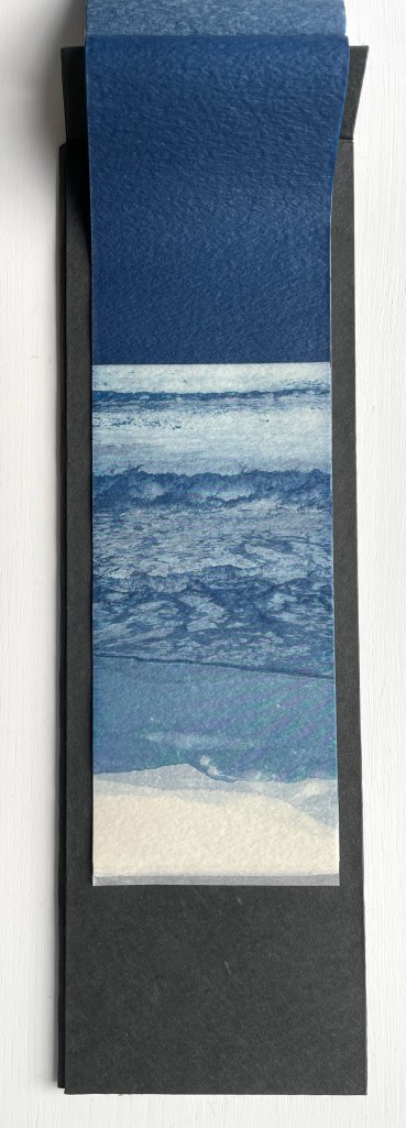 A bookmark featuring a watercolor design of ocean waves and sandy shore in shades of blue and white, resting on a black background.