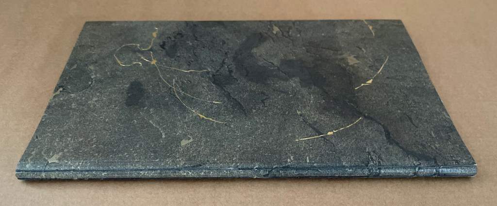 The cover of the book 'Amorous Embrace' by Suzanne Moore, made from stone with gold leaf accents, resting on a brown background.