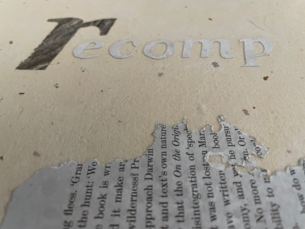 Close-up view of the word 'recomp' printed in white on a textured surface, with a piece of torn paper containing text partially visible below.