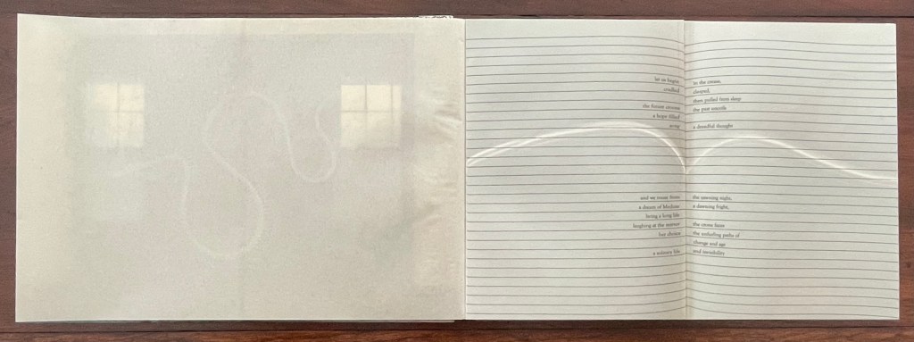 Open artist's book displayed on a wooden surface, revealing a gatefold spread. The left side shows a faint reflection of two windows, while the right side features lined pages with text arranged in a poetic format.