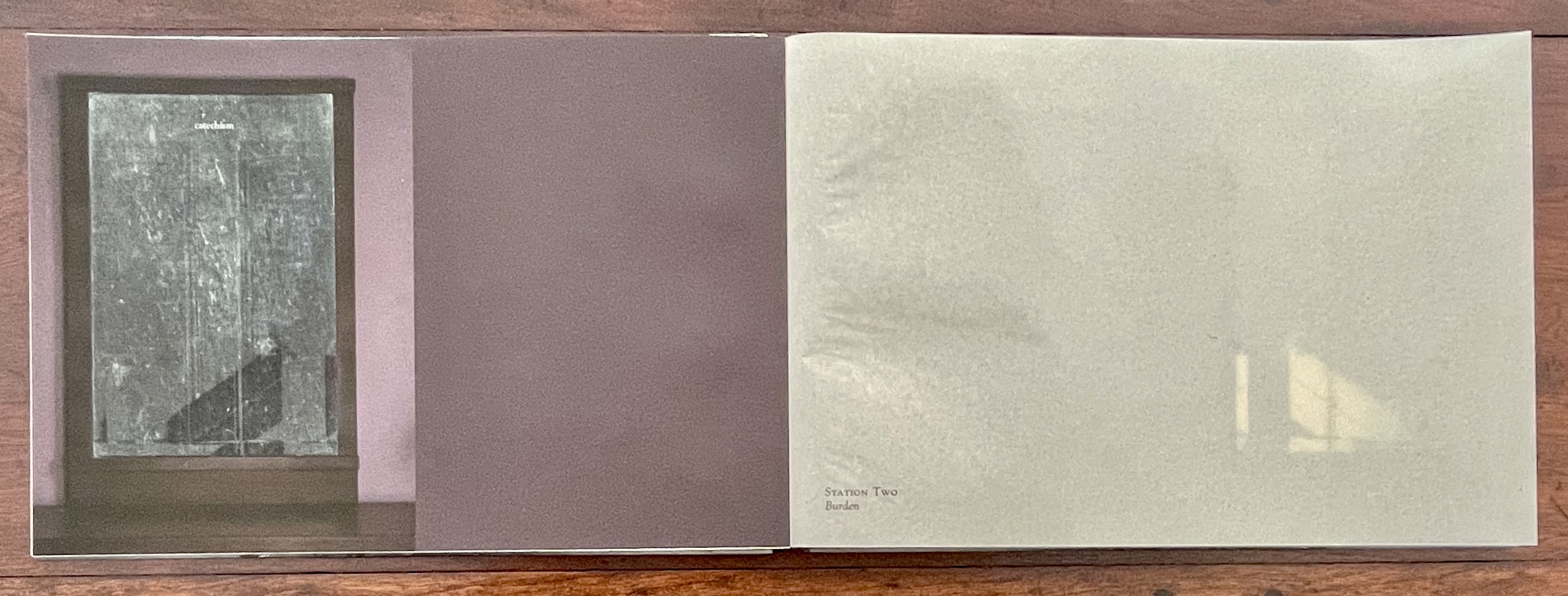 An open artist's book featuring a visible page with the word 'catechism' at the top, surrounded by a textured surface that appears to be a window frame adjacent to a smooth, monochromatic page on the right.