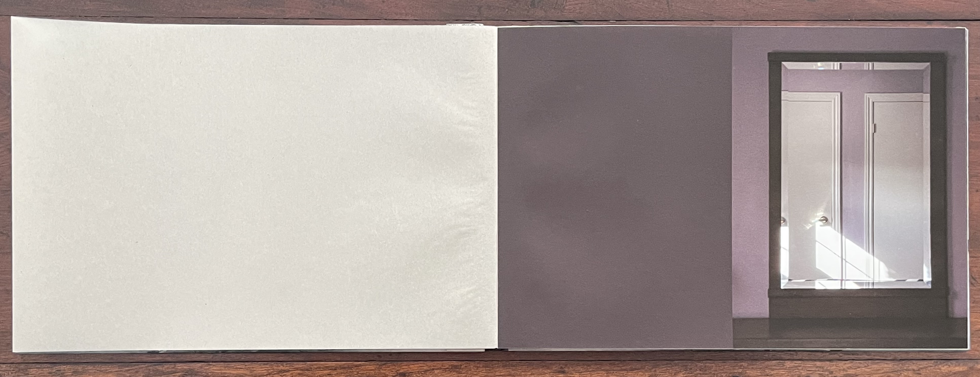 An open artist's book with a soft, blank left page and a right page displaying a photograph of a room featuring two closed doors and light casting shadows across the floor.
