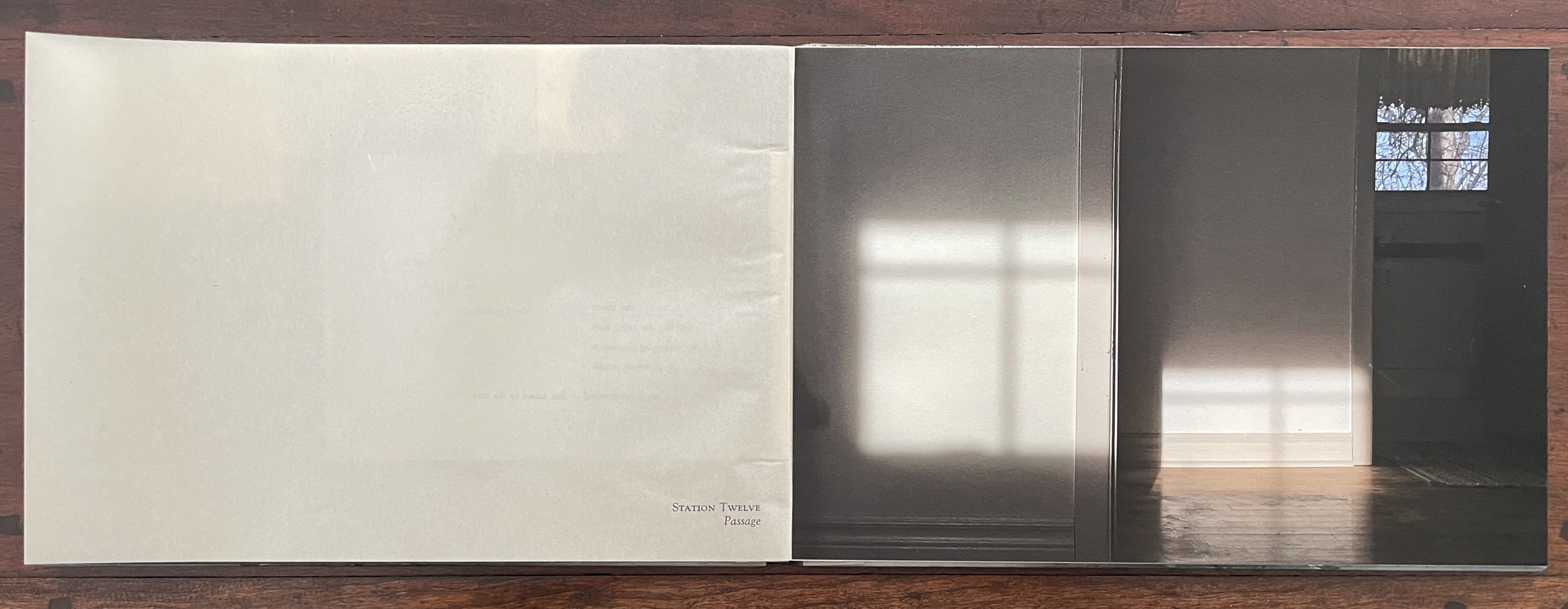 An open artist's book displaying a blank left page and a right page featuring a photograph of a light-filled room, revealing its shadow on the floor and a window with trees outside.