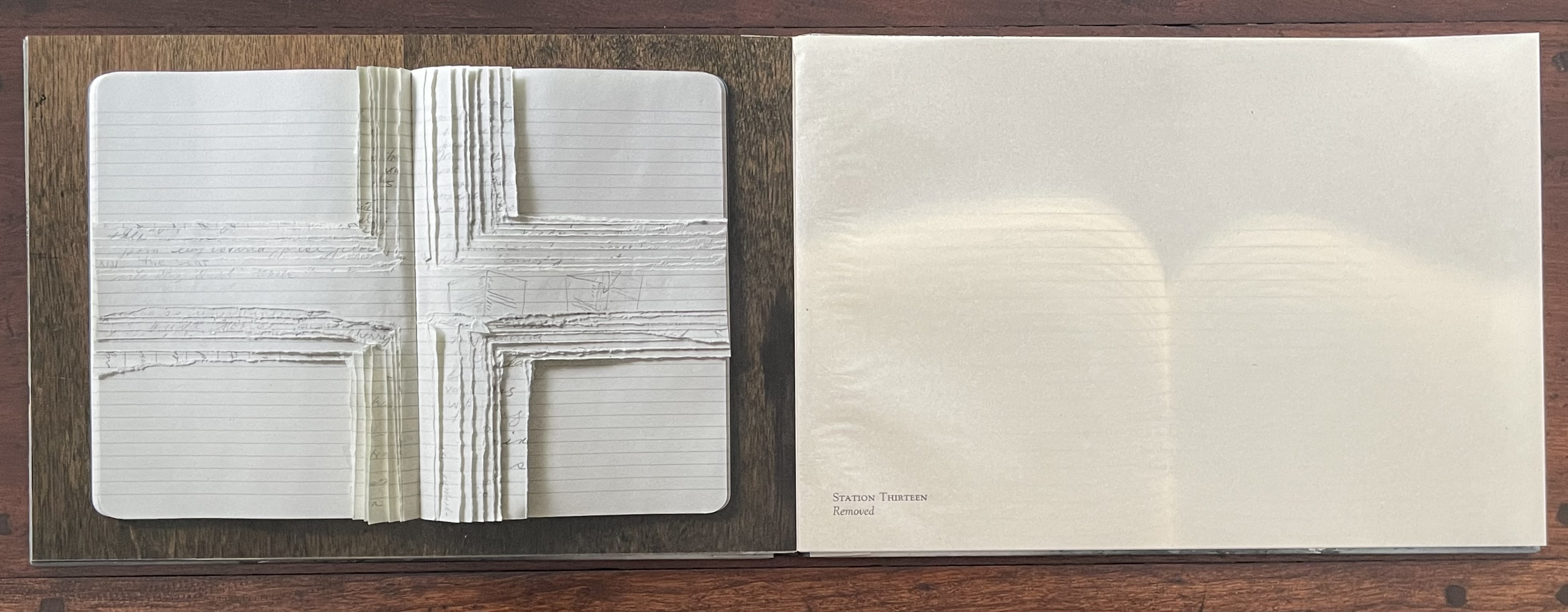 An open artist's book featuring layered pages arranged to form a cross shape, resting on a wooden surface with a smooth blank page overlapping on the right side.
