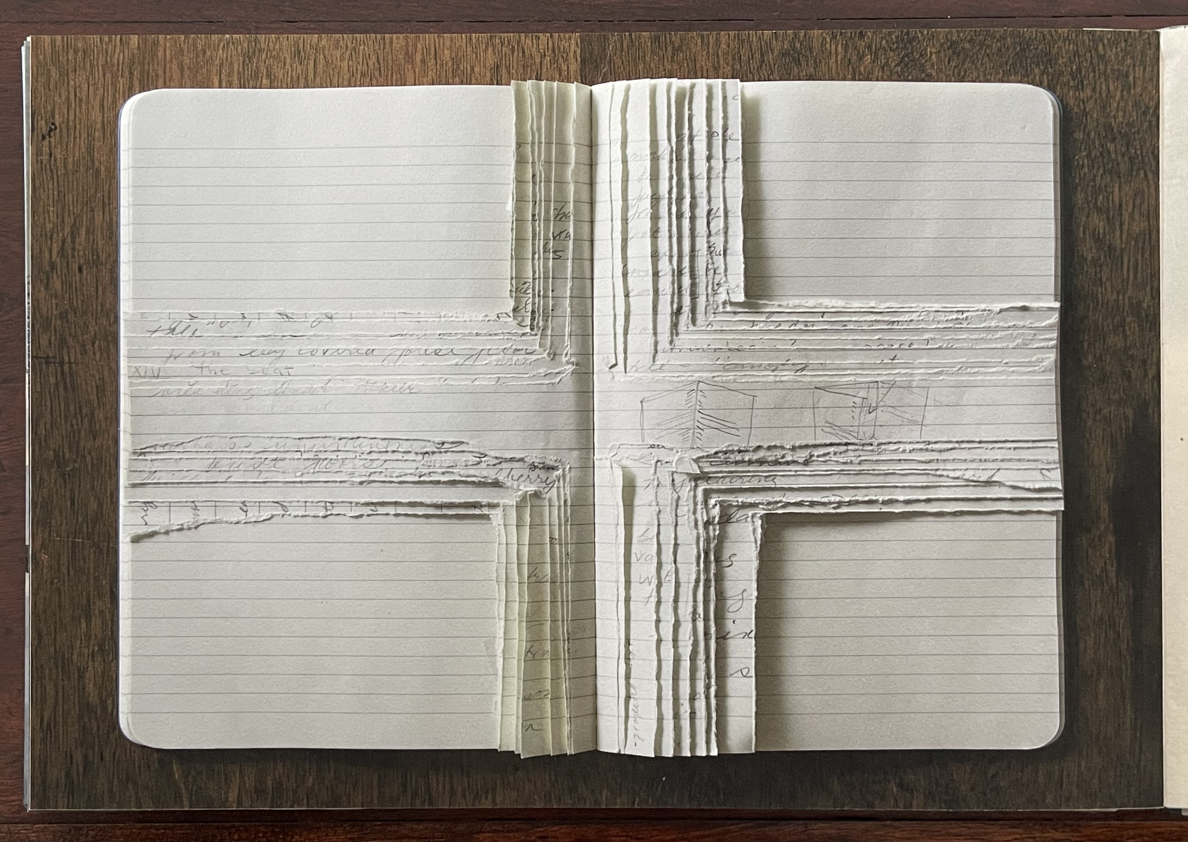 Open artist's book with a layered, textured design resembling a cross, featuring handwritten notes and lined pages, set against a wooden table background.