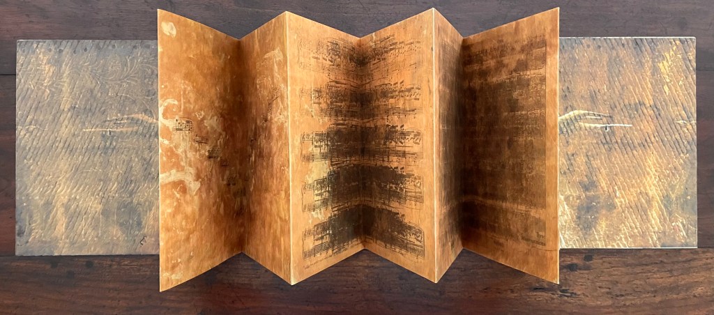 An open artist's book displayed flat on a wooden surface, showcasing an accordion fold structure with intricate printed designs and textures on the pages.