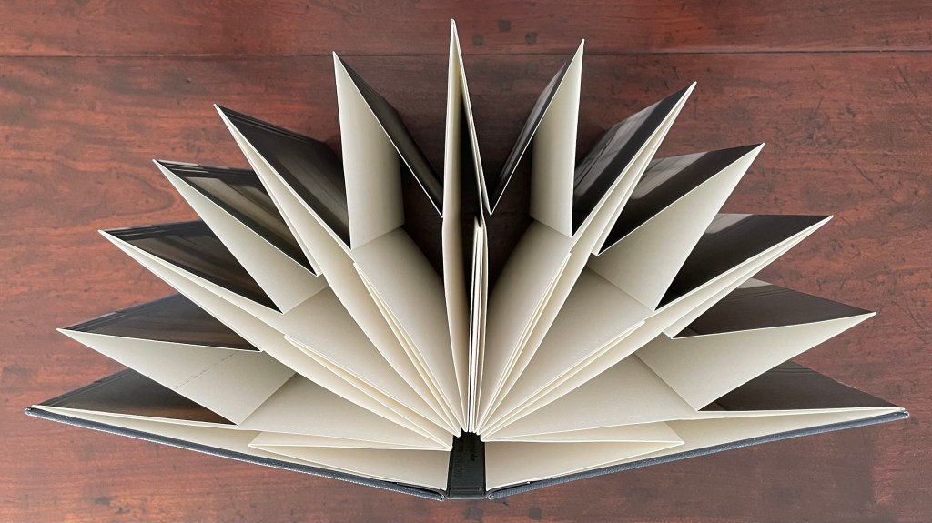 An open artist's book displaying an intricate accordion fold structure on a wooden surface.