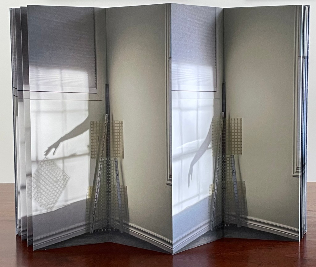 An artist's book displayed in a gatefold structure, showcasing layered images of a shadow against a wall, with a hand reaching toward an abstract object, possibly a sculptural piece or architectural model.