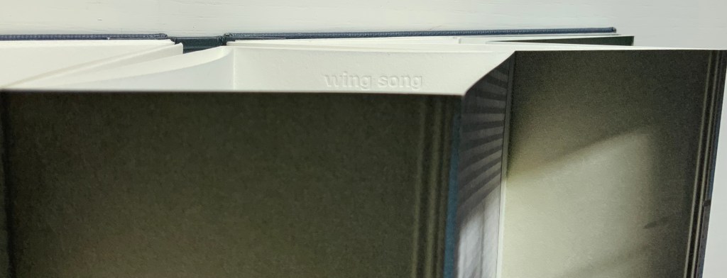 Close-up view of the inside binding of an artist's book by Marlene MacCallum, displaying the embossed title 'wing song' on the page edge against a contrasting dark interior.