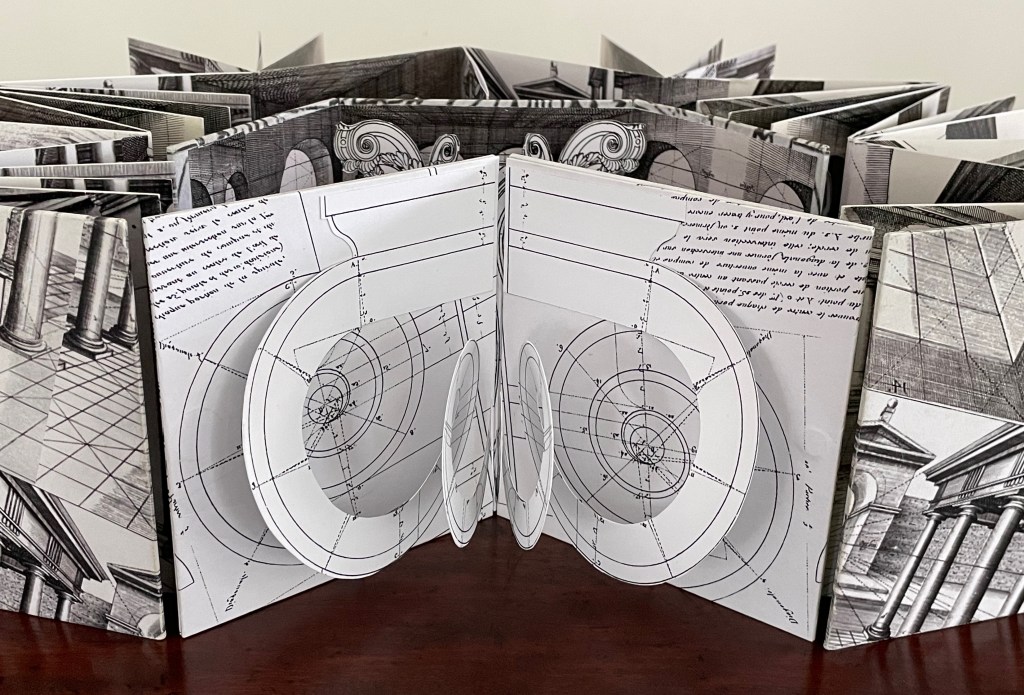 A detailed pop-up book featuring architectural diagrams and illustrations, with intricate folds and designs visible on multiple pages, displayed on a wooden surface.