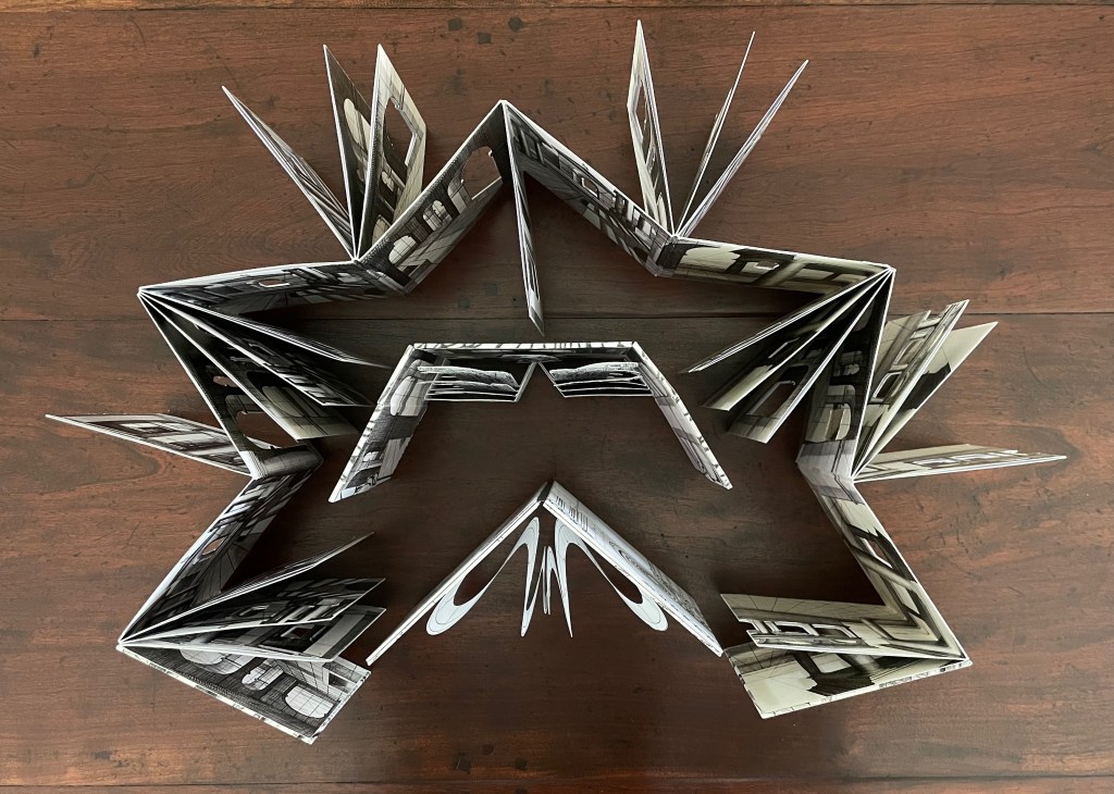 An artistic arrangement of a folded book displaying architectural photographs, shaped into a star-like form, placed on a wooden surface.