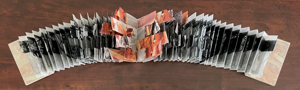 An open artist's book featuring a series of layered photographs in shades of black, white, and orange, arranged in a folding format on a wooden table.