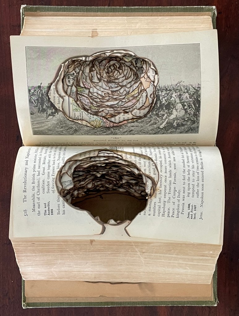 An altered book sculpture featuring a carved floral design on pages, showcasing an intricate layering and burning technique, with an illustration visible above.