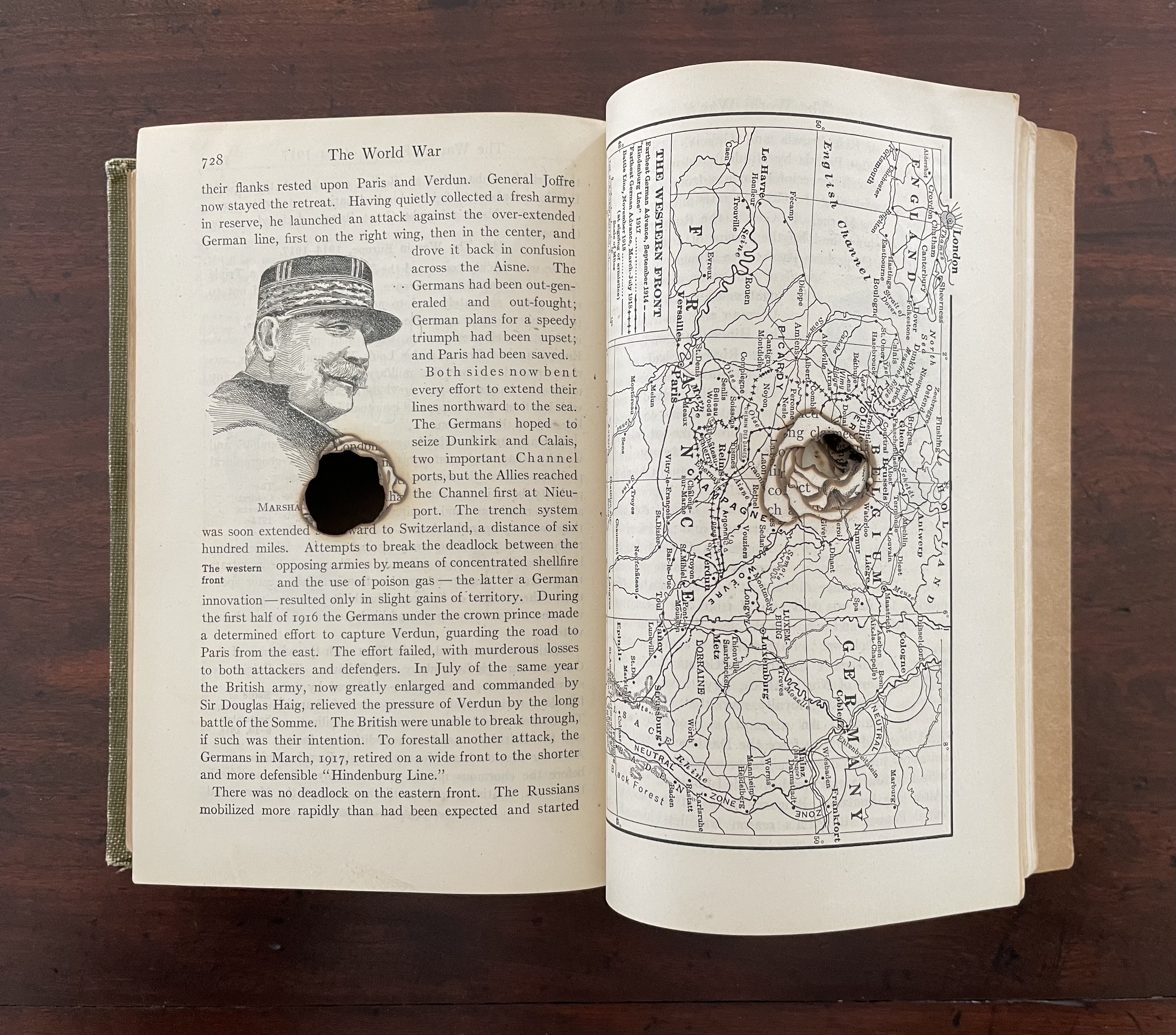 An open altered book featuring a page on World War I with an illustration of a soldier's profile and a detailed map, displaying a significant hole burned through the center.