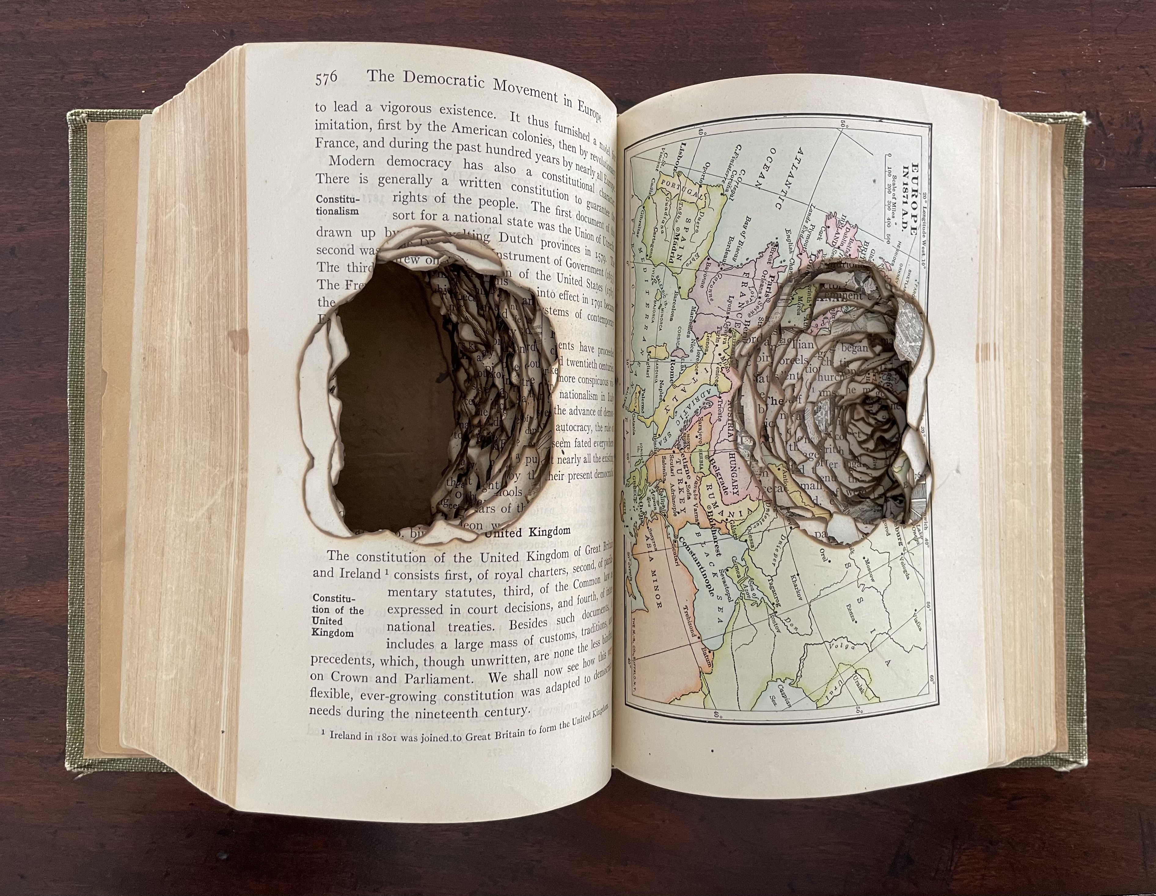 An altered book displaying a cut-out design, revealing intricate layers and shapes, positioned across two pages showcasing a map and text about democracy.