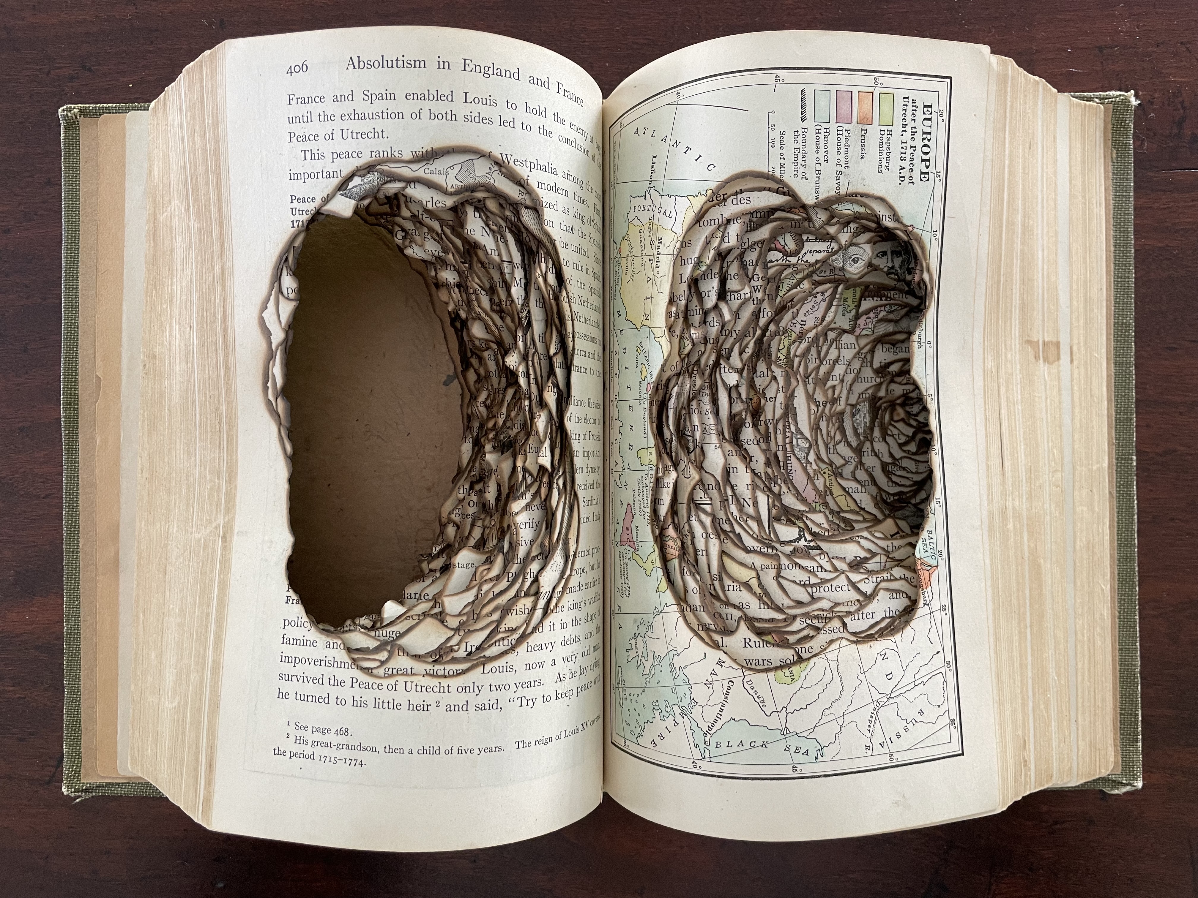 An altered book featuring two large, carved hollowed areas in the pages, surrounded by intricate, burnt, spiraling patterns, showcasing artistic reinterpretation of text and images.