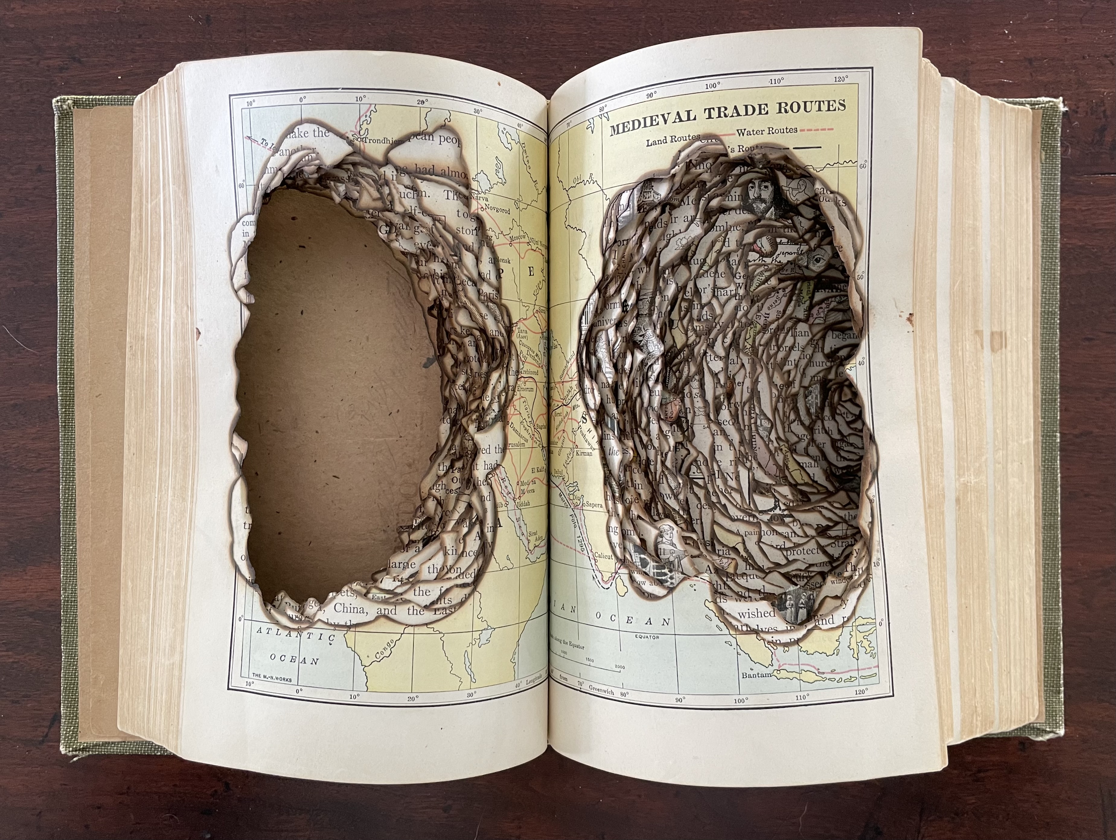 Open altered book showcasing a page with intricate pyrographic designs and cutouts highlighting medieval trade routes, revealing a textured layered effect.