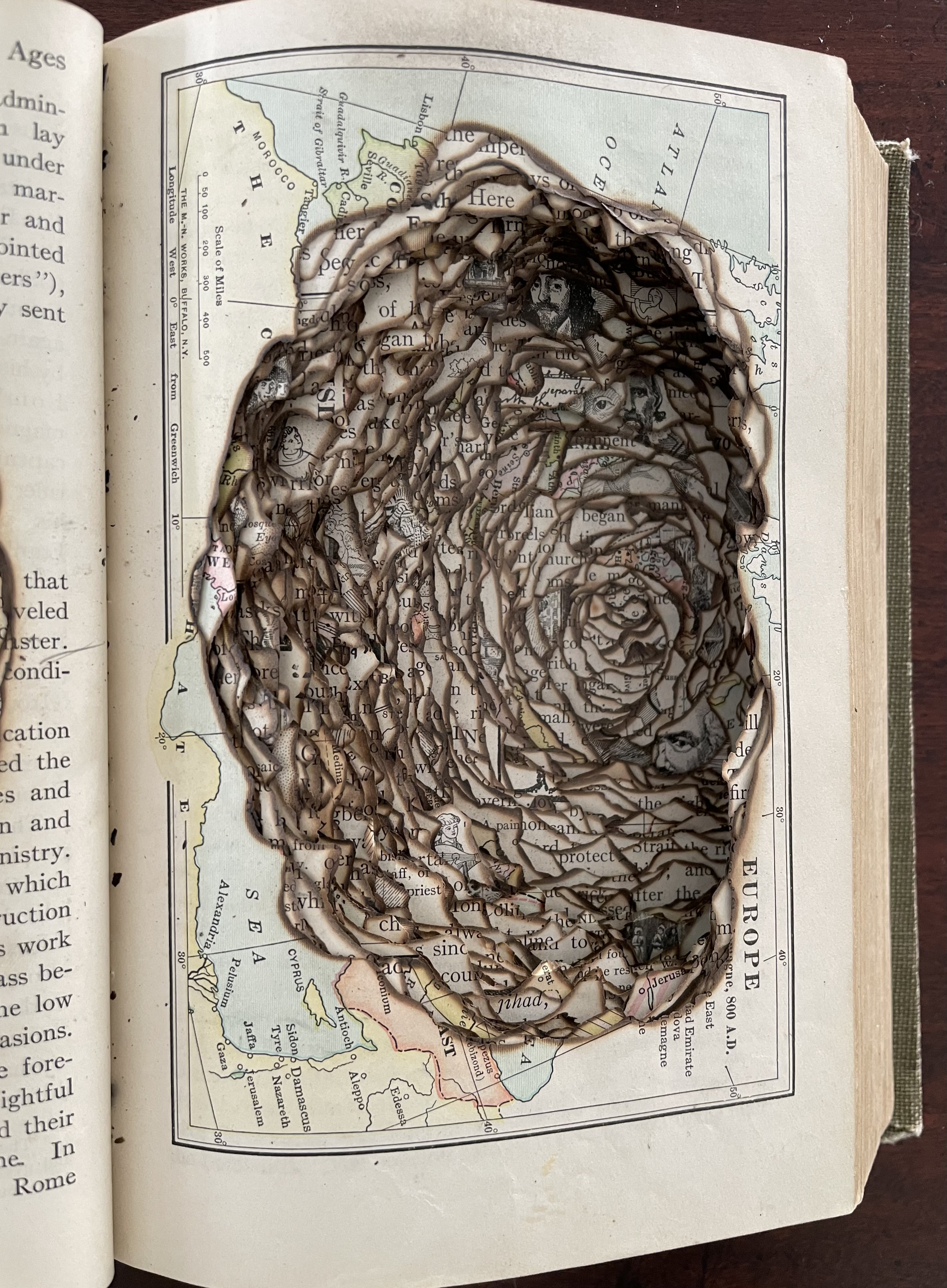 An altered book featuring a burned and sculpted section revealing layers of text and illustrations, showcasing artistic manipulation of historical maps.