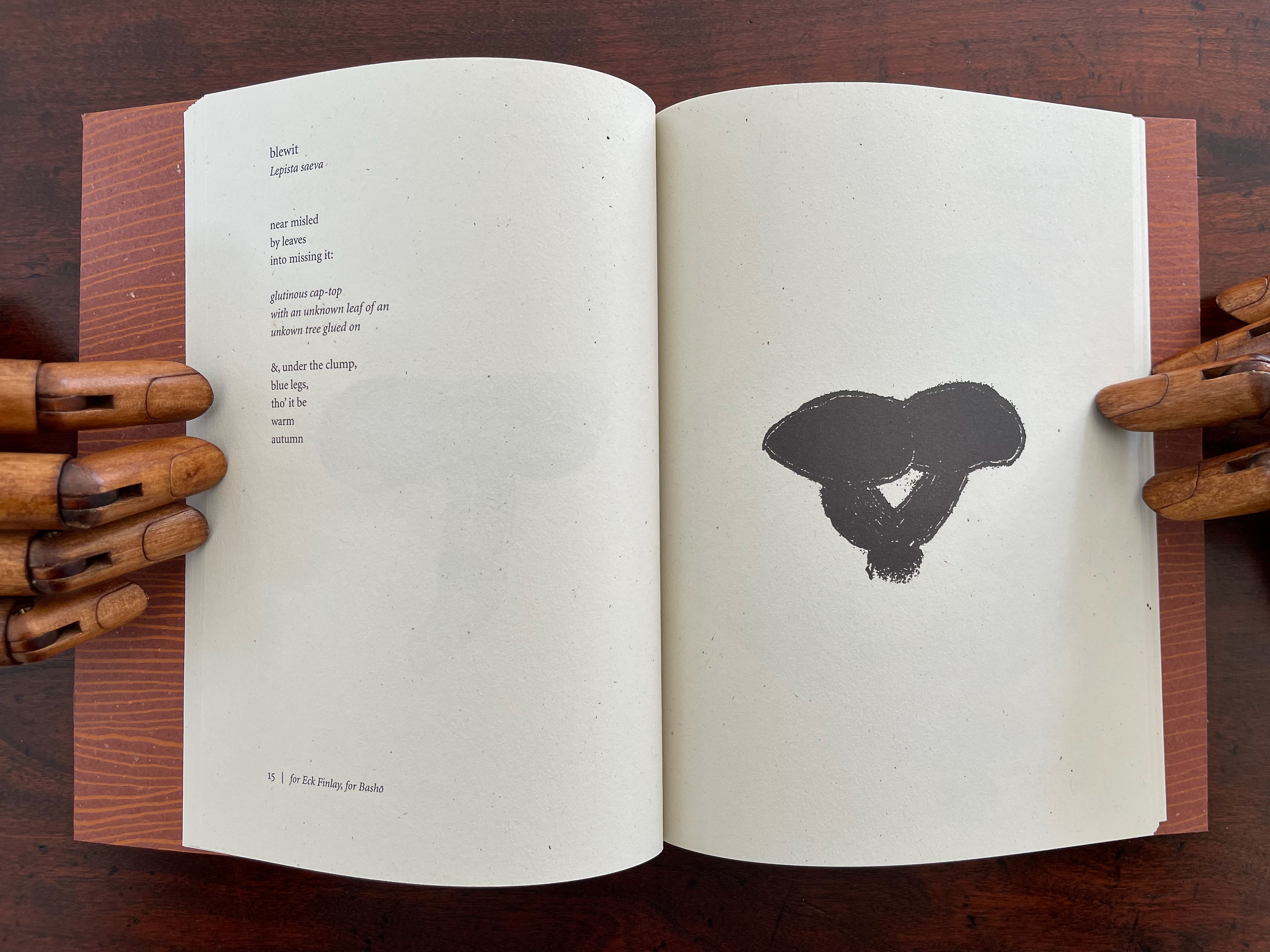 Open book displaying a poem titled 'blewit' on the left page, accompanied by a black ink illustration of a mushroom on the right page. The book rests on a wooden surface, held by carved wooden hands.