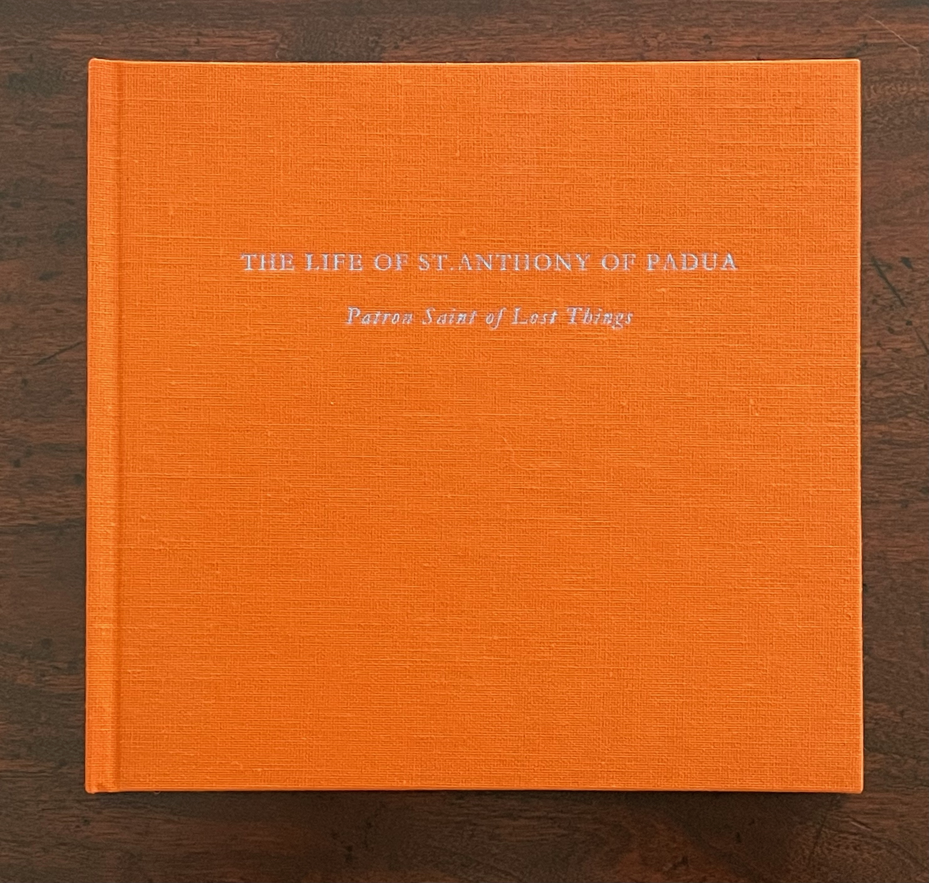 An orange hardcover book titled 'The Life of St. Anthony of Padua: Patron Saint of Lost Things'. The cover features silver text and a textured finish.