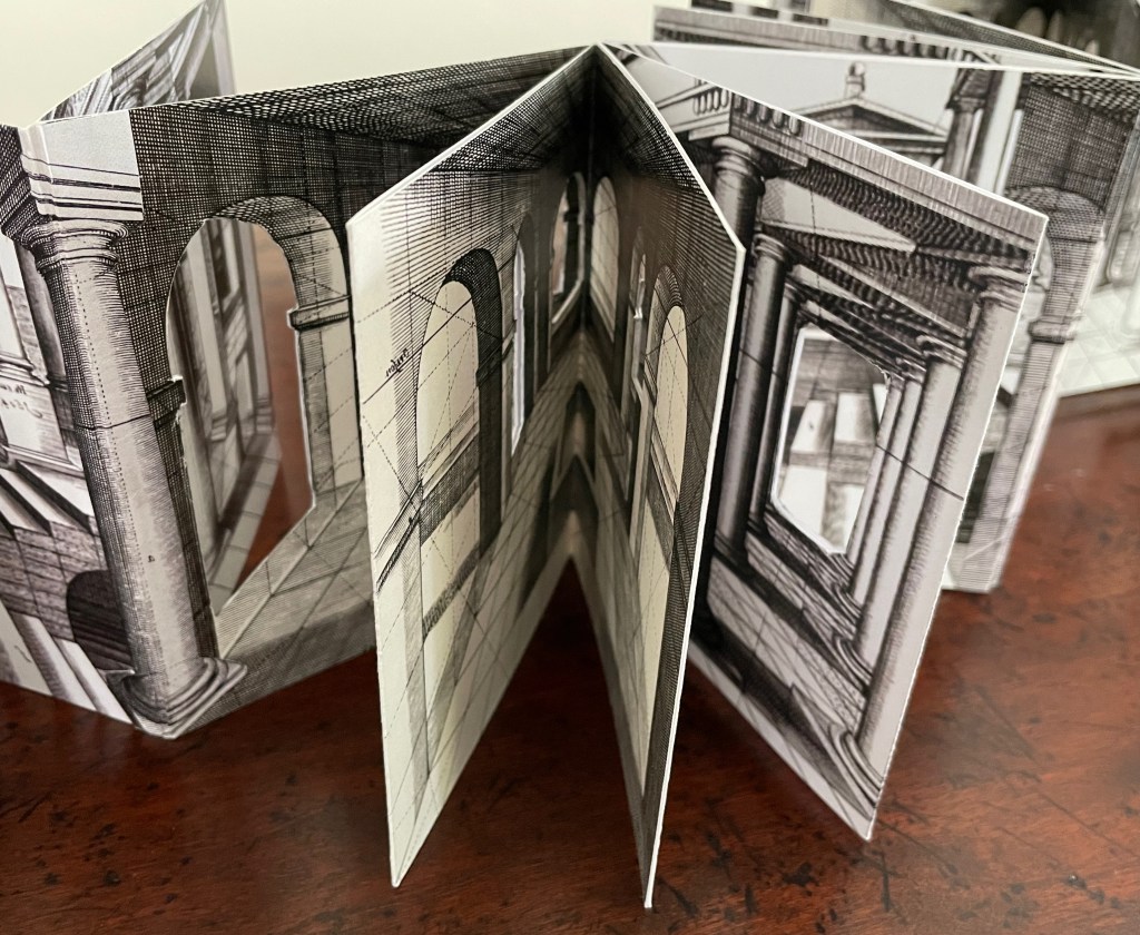 An open artist's book featuring intricate black-and-white architectural illustrations, displaying columns and arches in a zigzag format on a wooden surface.