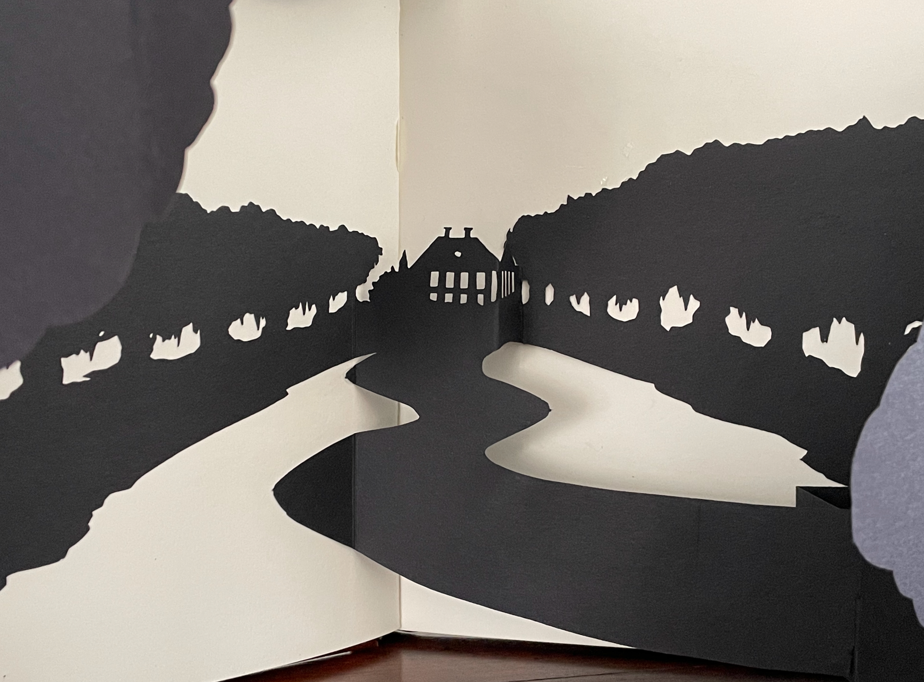 A black paper cutout scene depicting a winding road leading to a house, surrounded by silhouettes of trees.