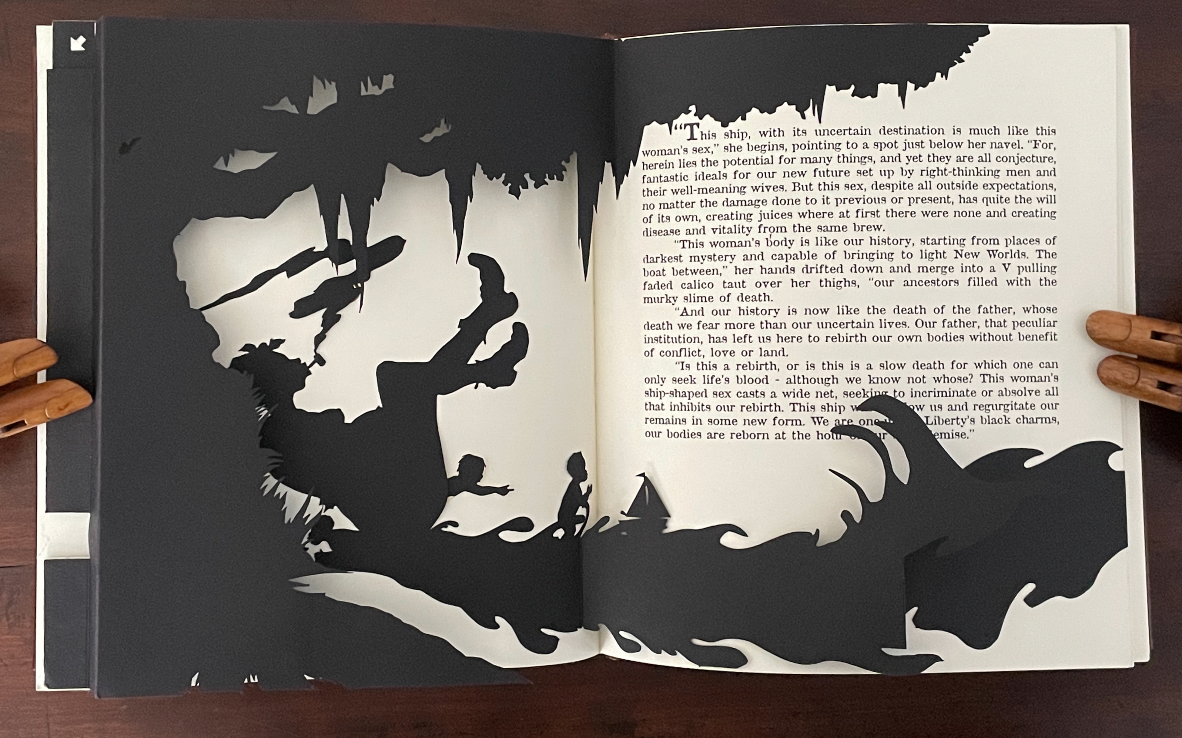 An open book with layered black cutouts creating a silhouette of figures and a ship against a white background, accompanied by text on the right page discussing themes of rebirth and mystery.