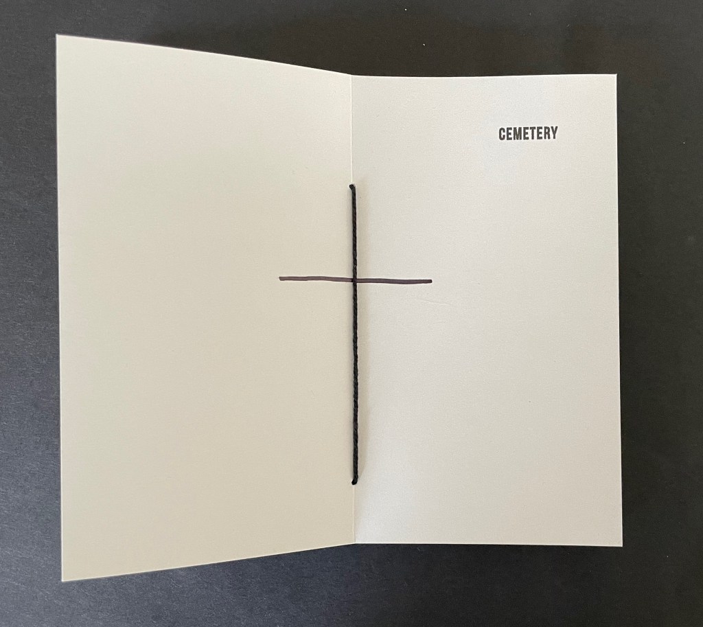 An open booklet with the word 'CEMETERY' printed on the right page, and a simple cross drawn in brown ink across the center.