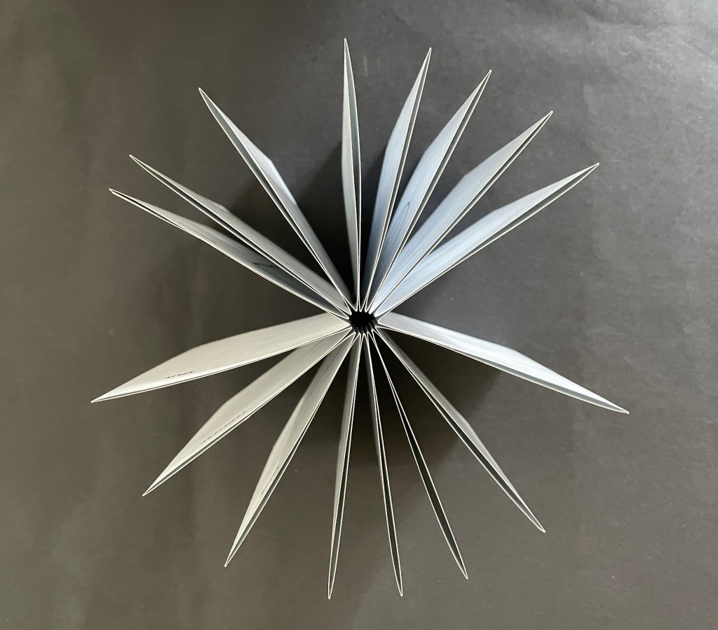 An overhead view of an open book with multiple pages fanned out in a star-like arrangement against a dark background.