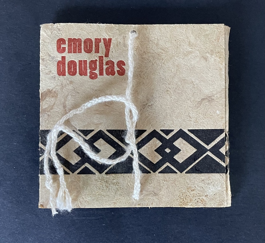 Close-up of a small, handmade book with the name 'Emory Douglas' printed in red and a decorative black pattern at the bottom. The book is tied with a piece of string.