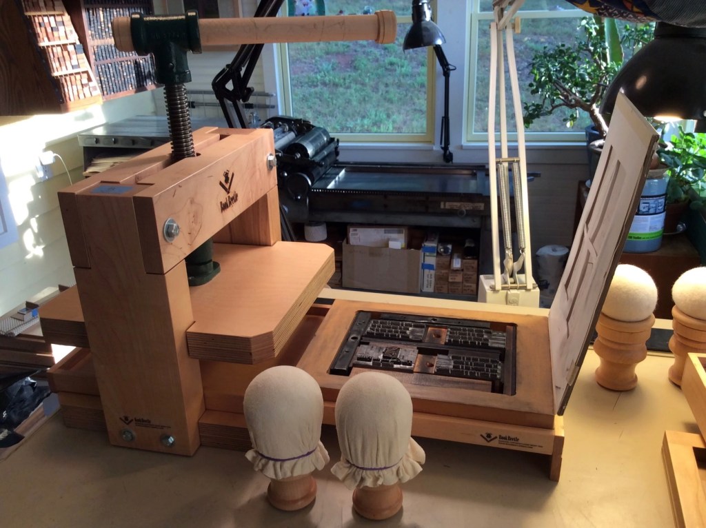 A wooden book press positioned on a workspace, featuring a large handle, a platform for printing materials, and two covered objects resembling human figures, with natural light illuminating the scene.