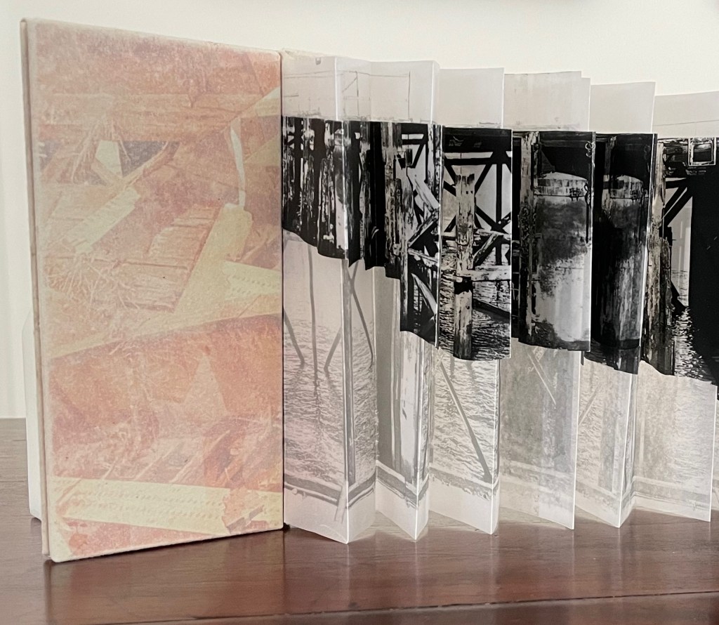 An art book with a textured cover in muted colors, fanned open to reveal a series of black and white photographs printed on translucent pages, depicting wooden structures and water reflections.