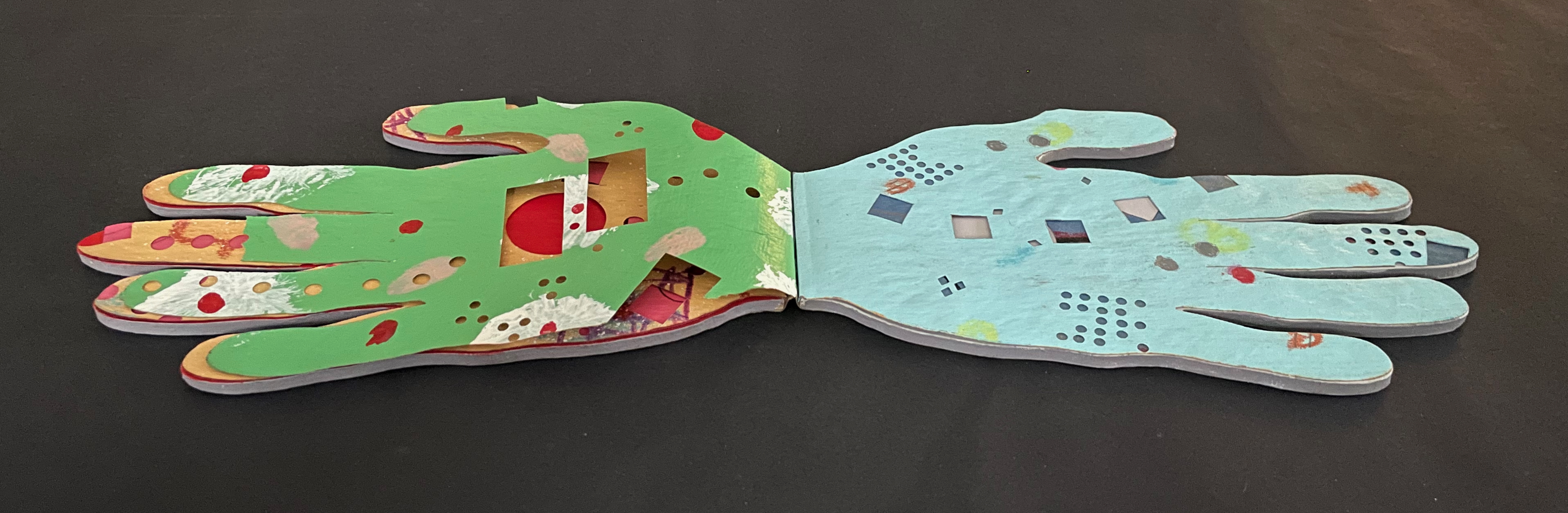 An open book with two hand-shaped pages, one side painted green with colorful patterns and cutouts, the other side painted blue with geometric shapes and dots.