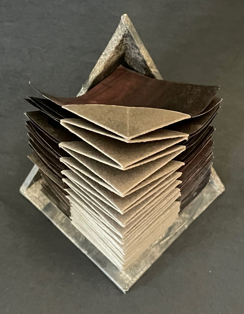A geometric sculpture made of stacked square pages in varying shades of brown and gray, forming a pyramidal shape with a solid base.