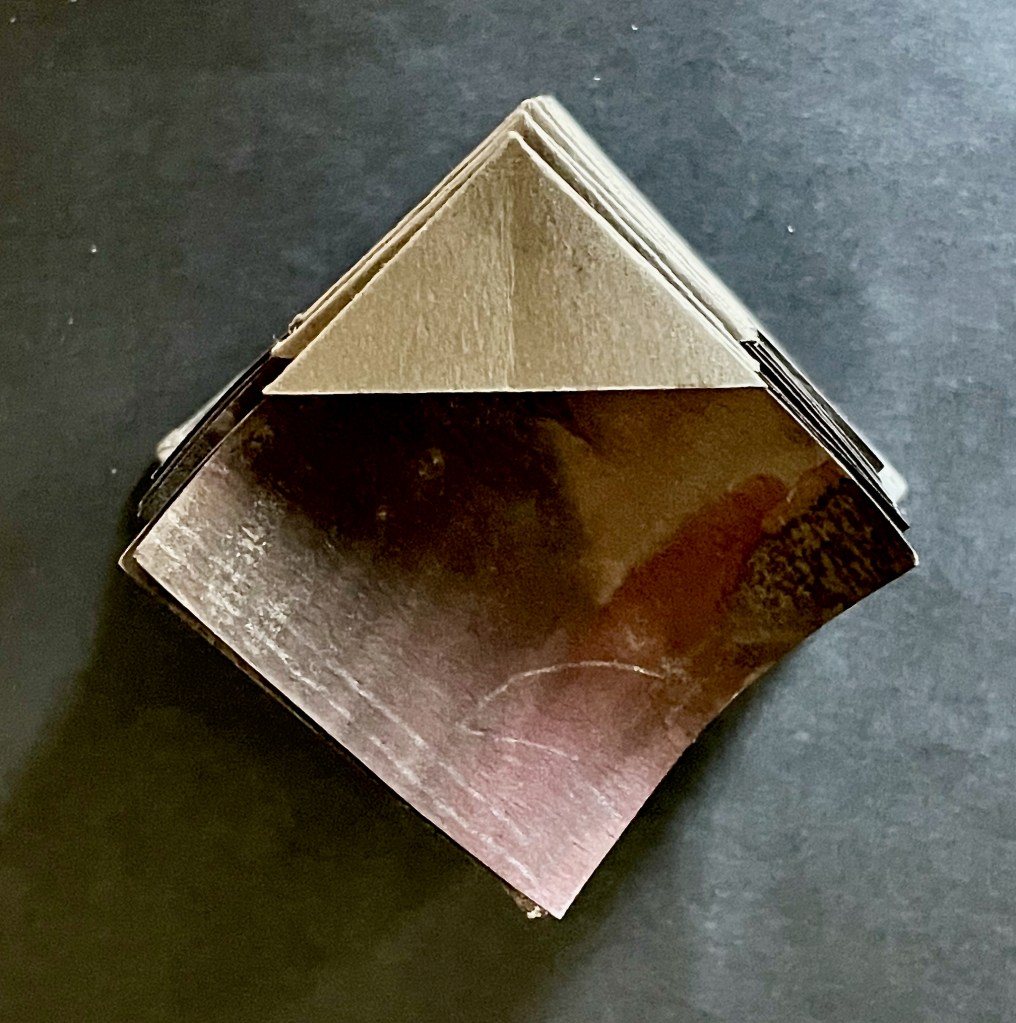 A top view of a paper object resembling a miniature pyramid, made from folded paper with a glossy texture on one side, resting on a dark surface.