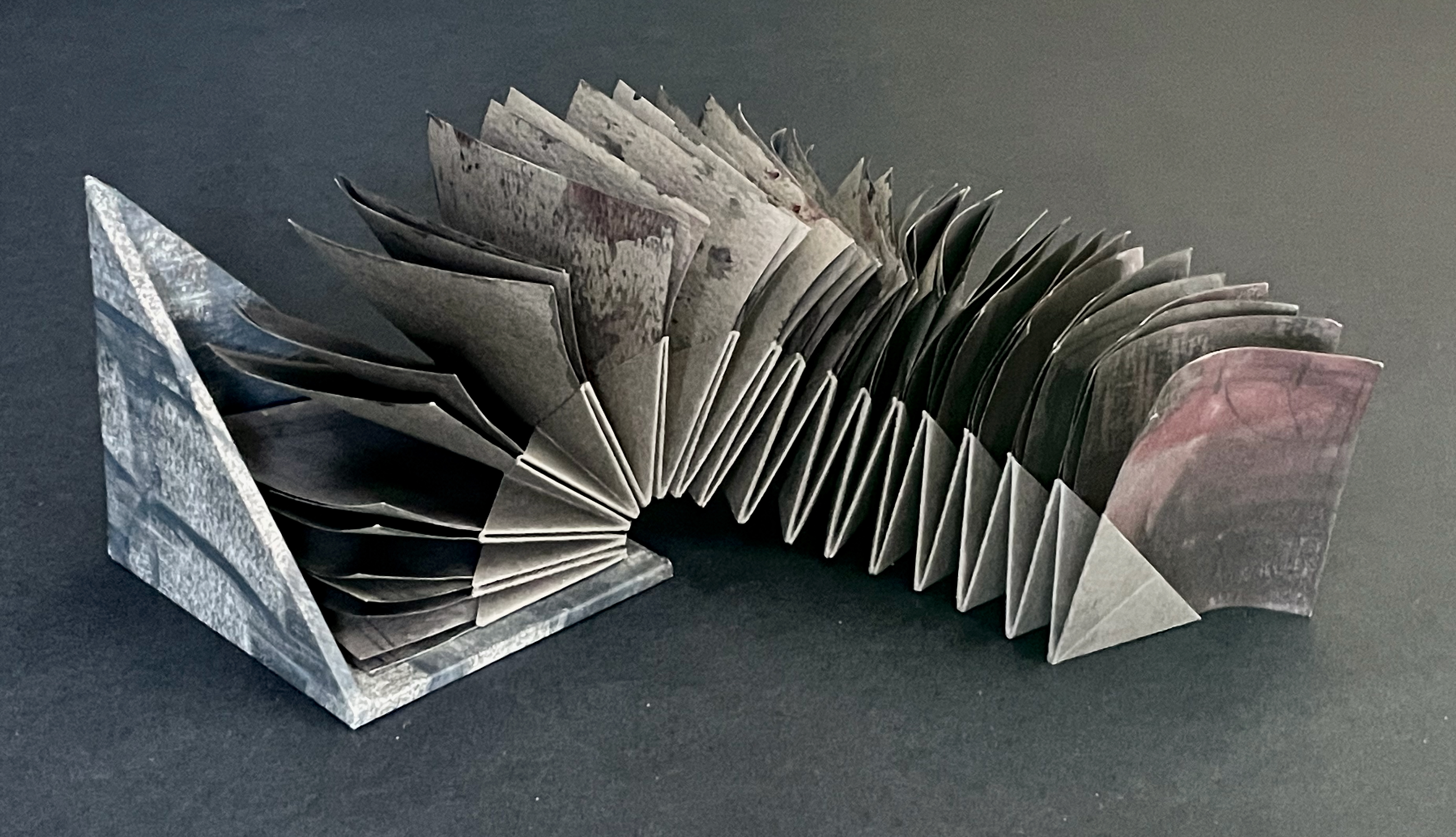 A sculptural book with a zigzag design, featuring textured pages in shades of gray and purple, resting on a dark surface.
