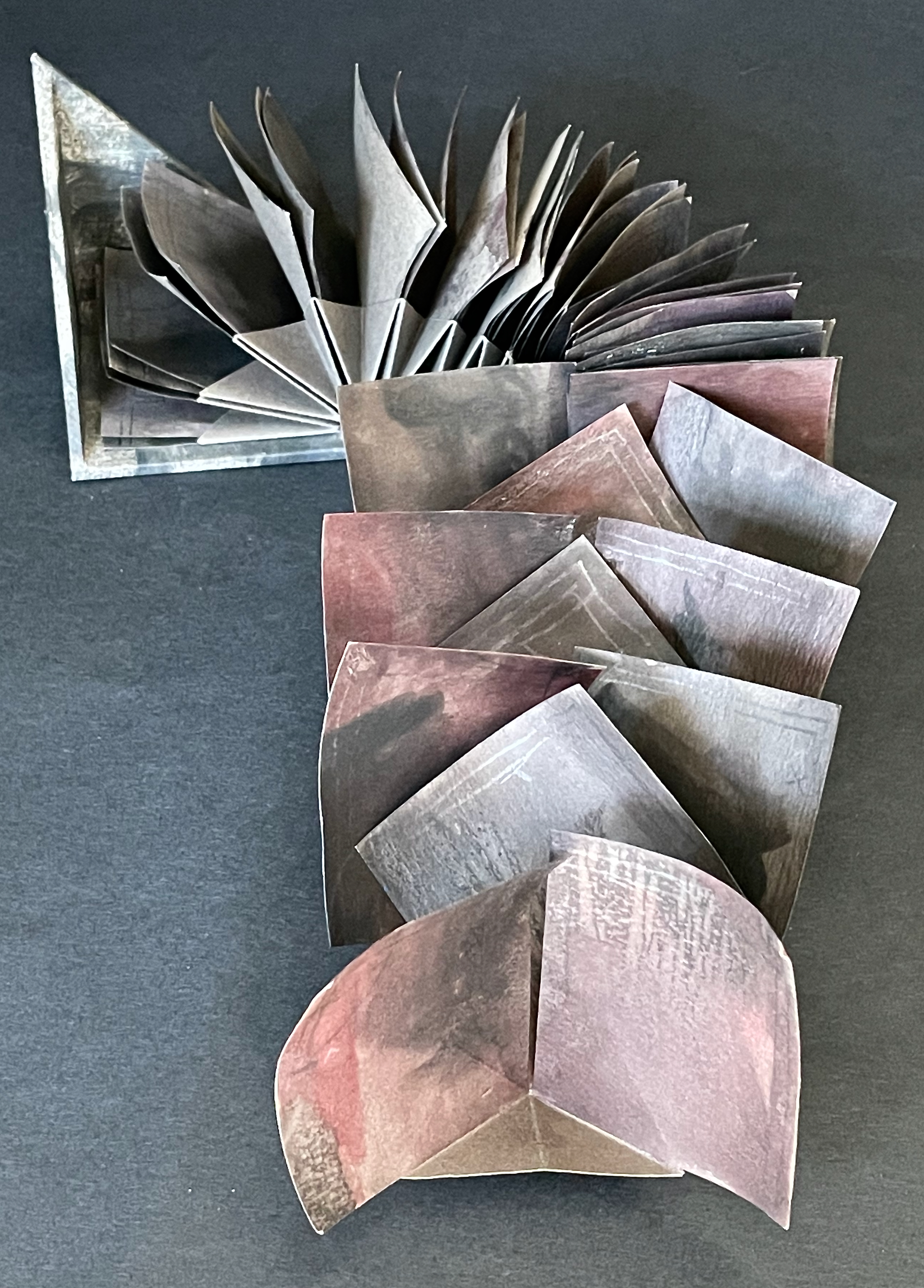 An artistic arrangement of layered, folded paper pages in varying shades of gray, pink, and brown, fanned out and partially stacked.