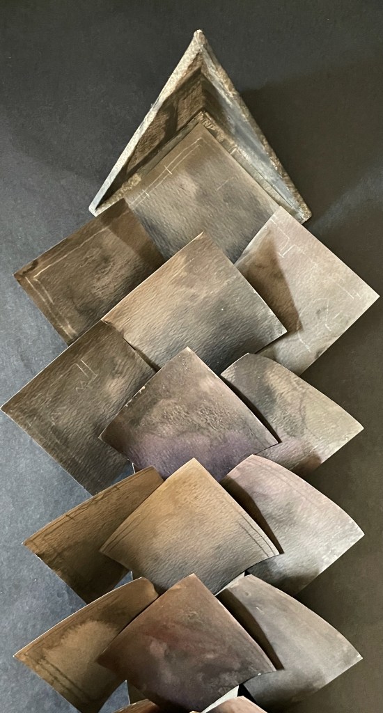 An artistic arrangement of layered, curved metal pieces forming a pyramid-like structure against a dark background.