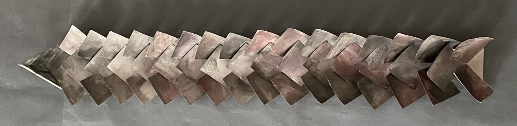 A series of abstract, curved metal shapes arranged in a zigzag pattern, showcasing a gradient of muted colors from gray to bronze against a dark background.