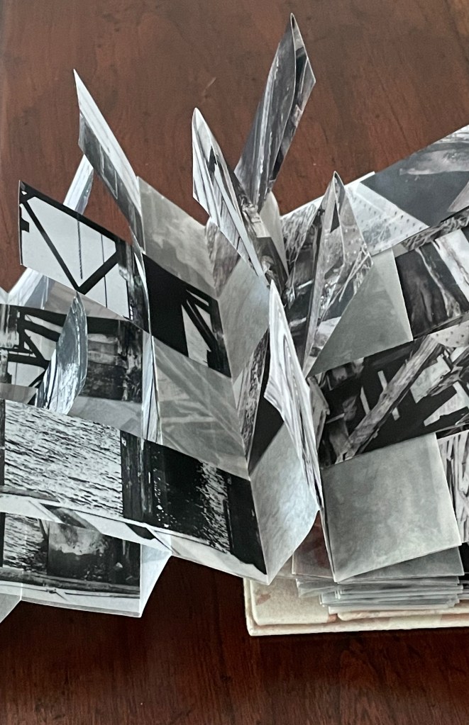 A pop-up book structure made of black and white photographs, displaying various images that extend outward. The pages are intricately arranged, creating a dynamic three-dimensional effect against a wooden surface.