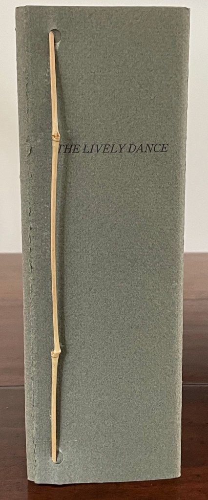 A vertical view of a grey booklet titled 'THE LIVELY DANCE' secured with a thin bamboo stick.