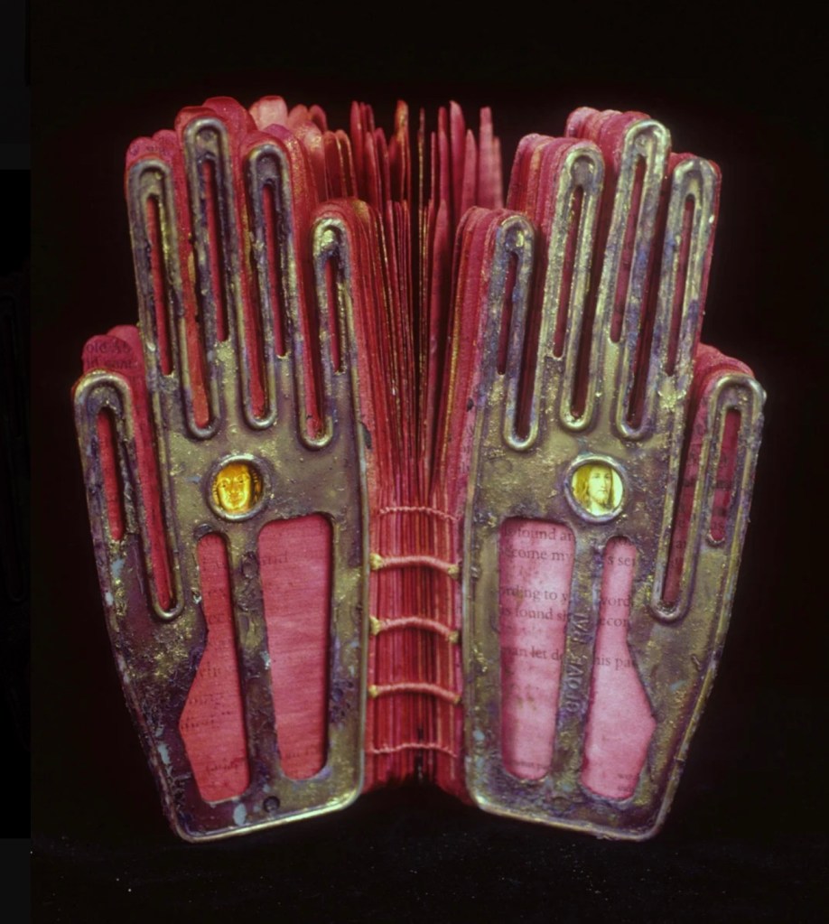 A unique book design featuring two metallic hand shapes as covers, with a red spine and numerous pages visible in between, some pages displaying text.