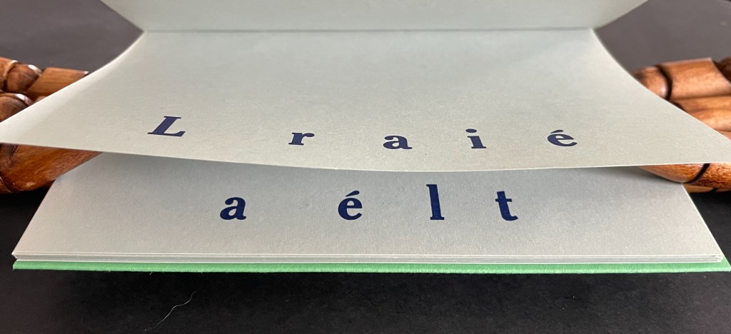 A partially opened book with letters printed on the pages, showcasing the text 'L raié a élt' on a light background, with a wooden object partially visible beside it.