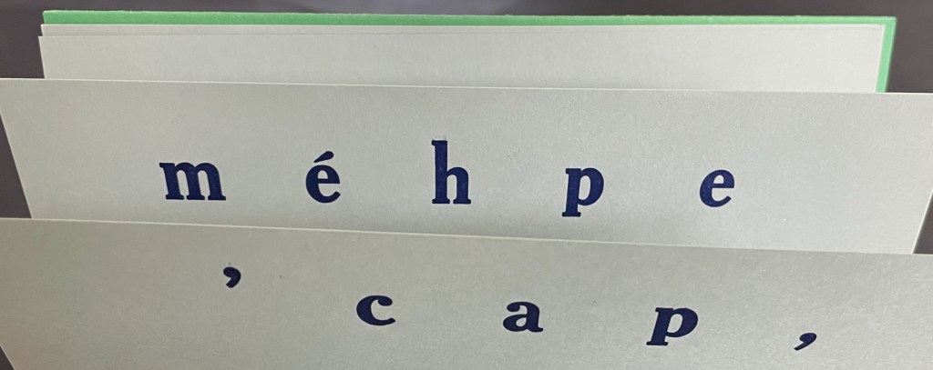 Close-up of a folded card with the words 'méhpe , cap,' printed in dark blue on light gray paper, with a green edge.