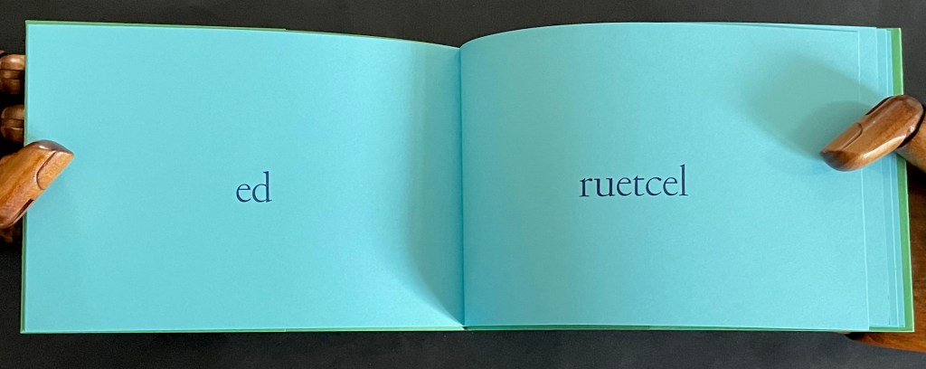 An open book with a turquoise background displaying the words 'ed' on the left page and 'ruetcel' on the right page, supported by wooden hands.