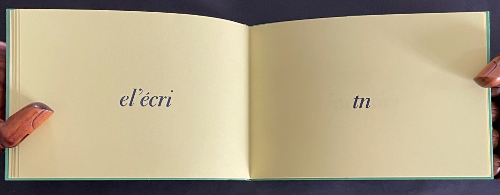 An open book with yellow pages displaying the words 'el'écri' on the left page and 'tn' on the right page, held by wooden hands.