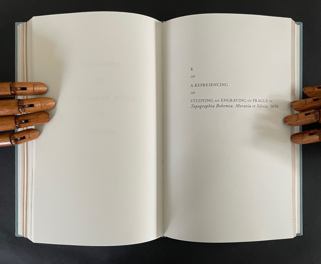 An open book with wooden mannequin hands holding the pages, displaying the title 'K or A Representing or Studying an Engraving of Prague in Topographia Bohemiae, Moravia et Silesiae, 1650' on the right page.